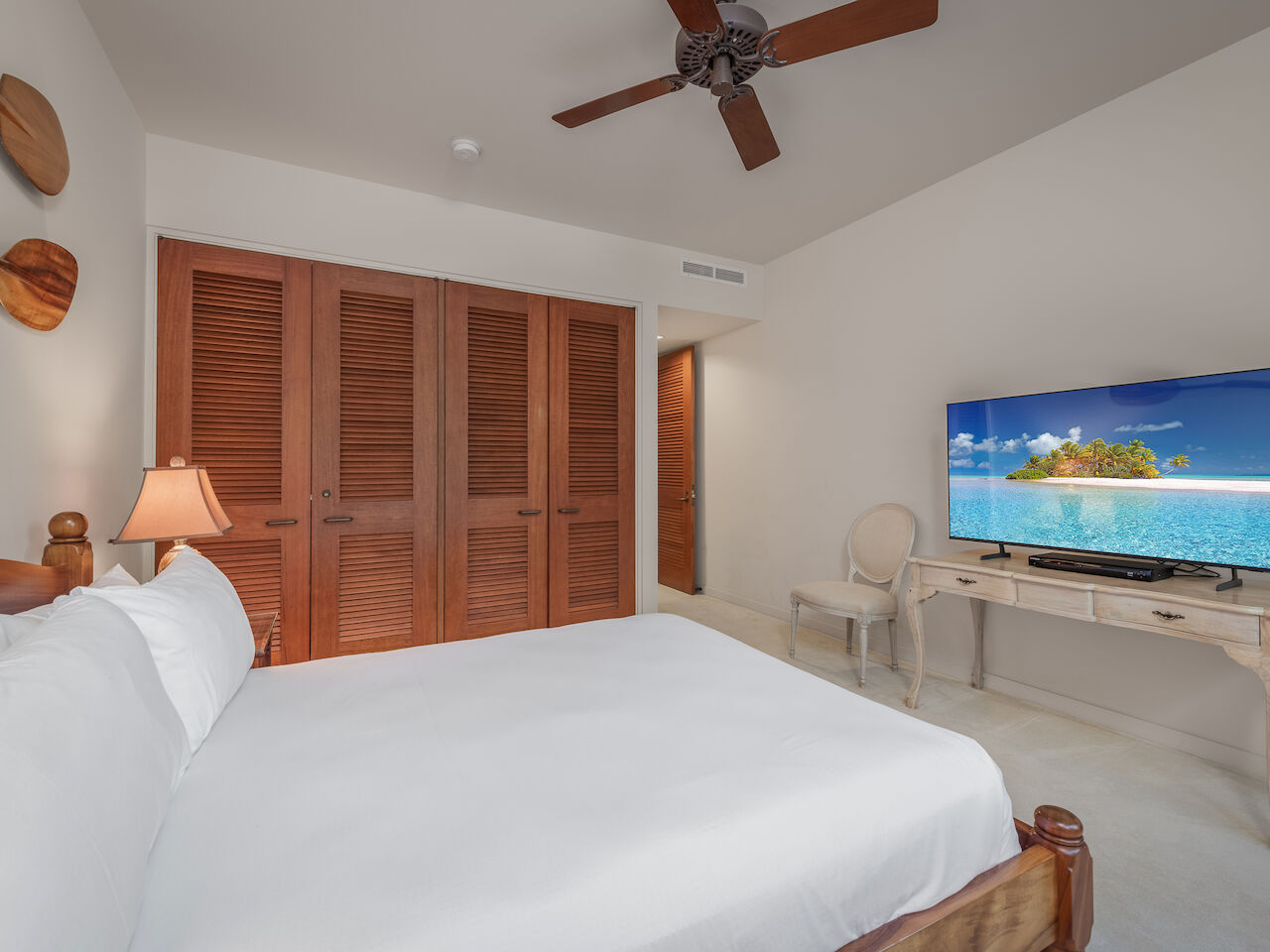 A bright beach-style bedroom with a white bed, wooden shutters, ceiling fan, closet doors, a desk with a TV, and light decor. end