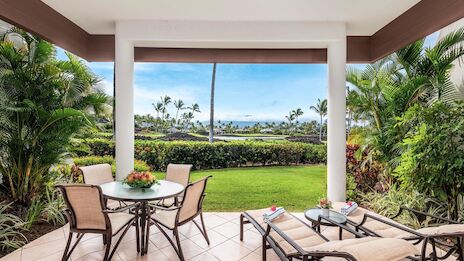 A covered patio with four chairs around a round table and a sunlit lawn beyond, lush tropical plants framing a peaceful garden view.