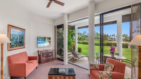 A bright living room with pink couches, a wooden coffee table, a TV, ceiling fan, and large glass doors opening to a green, sunny outdoor patio.