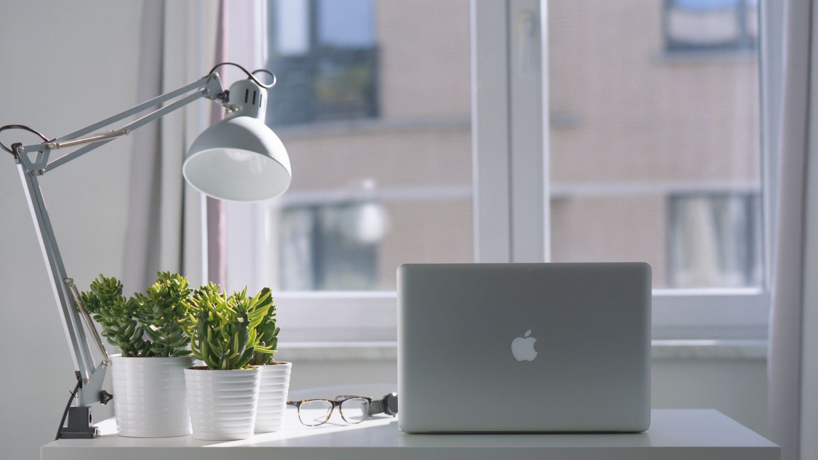 A minimalist desk setup with a laptop, desk lamp, glasses, and potted plants, set in front of a window with a view of buildings outside.