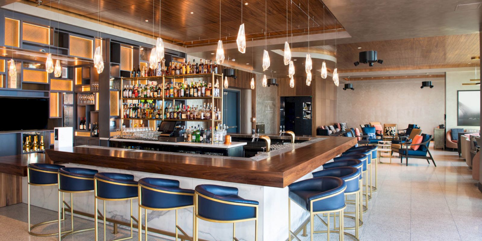 The image shows a modern bar with a wooden counter, stylish barstools, a display of various liquor bottles, pendant lights, and seating areas in the background.