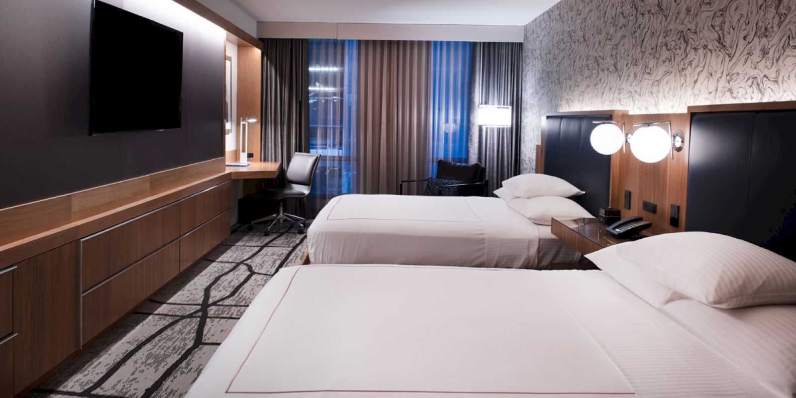 The image shows a modern hotel room with two beds, a desk, chair, TV, and lamps. The room has stylish decor with a gray and wood color scheme.