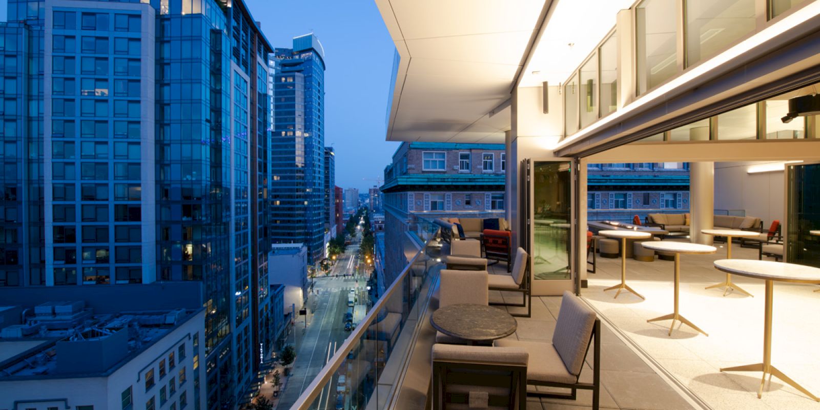 A modern, cityscape view from a high-rise balcony at dusk. The balcony has seating and tables, with buildings and a street visible below.
