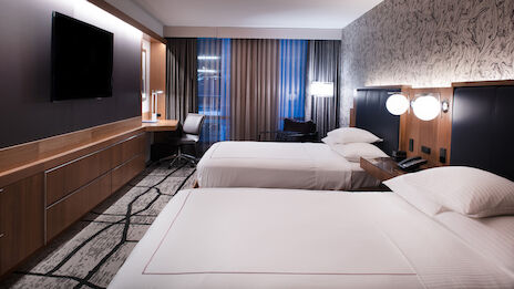 The image shows a modern hotel room with two neatly made beds, a flat-screen TV, a desk with a chair, and large windows with curtains.