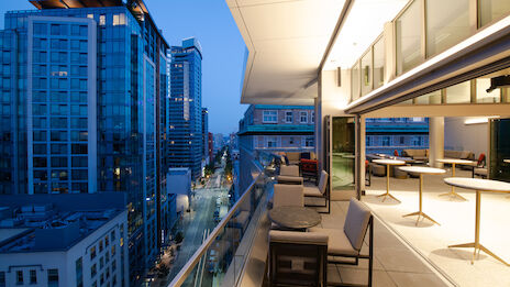 This image shows a modern cityscape at dusk with tall buildings and a well-furnished balcony extending from one of the buildings.