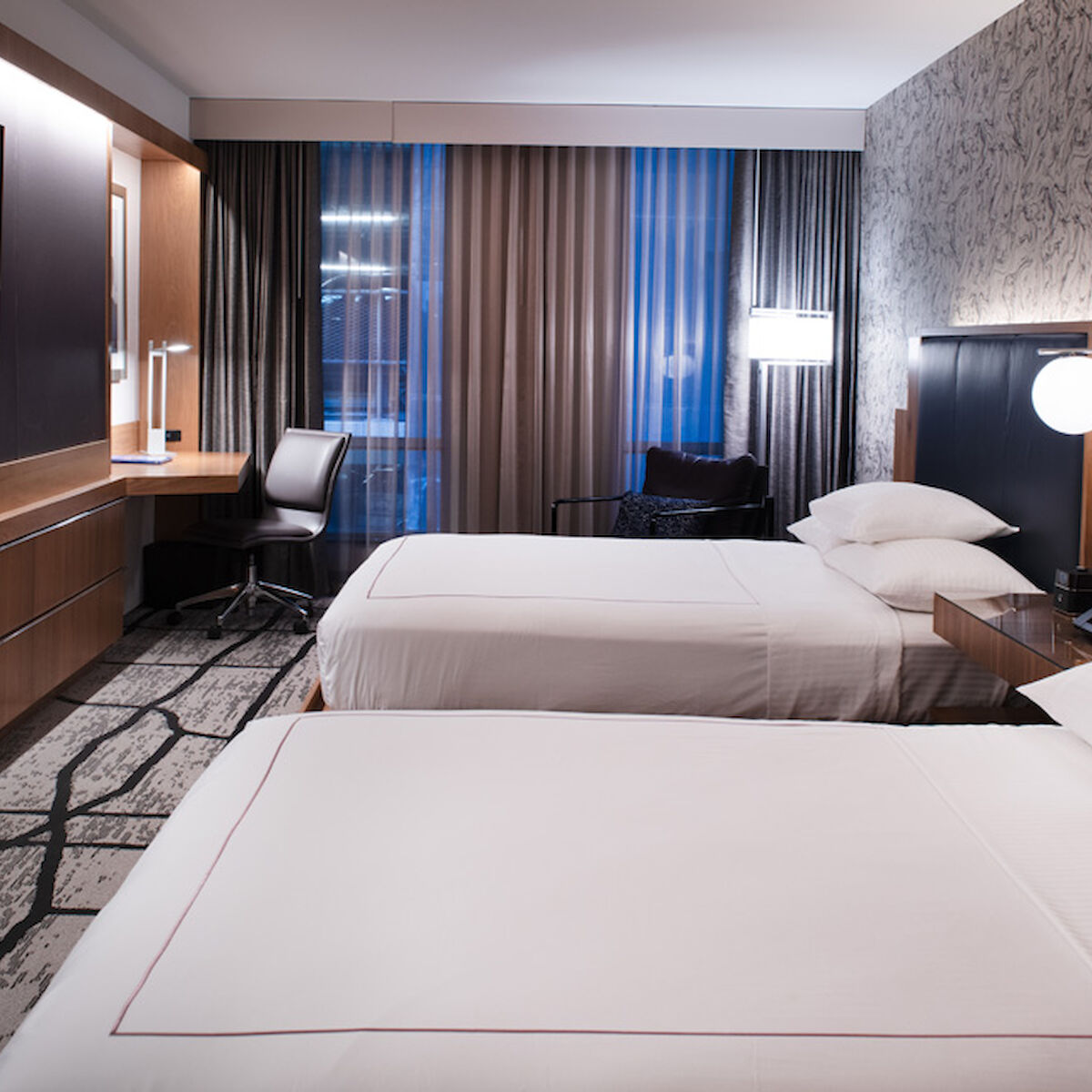 The image shows a modern hotel room with two double beds, a wall-mounted TV, a desk with a chair, and contemporary decor.
