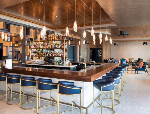 The image shows a modern bar with a wooden counter, elegant blue and gold bar stools, hanging pendant lights, and shelves stocked with various bottles.