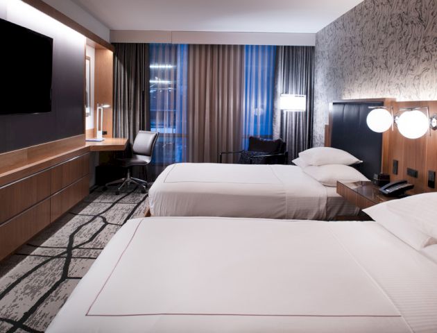 A modern hotel room with two double beds, a flat-screen TV, a desk with a chair, and a large window with curtains.
