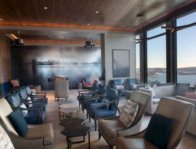 The image shows a modern, spacious lounge with large windows overlooking a scenic view, featuring ample seating and a mural of a city skyline on one wall.