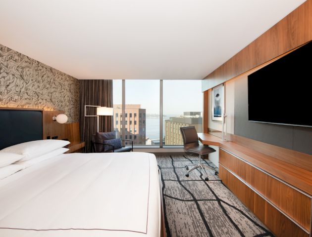 A modern hotel room with a large bed, desk, chair, and flat-screen TV. The window offers a cityscape view.