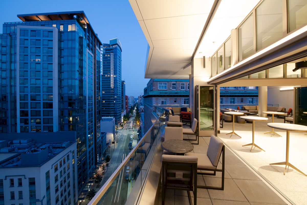A modern cityscape view at dusk from a high balcony, with seating areas and glass buildings in the background.