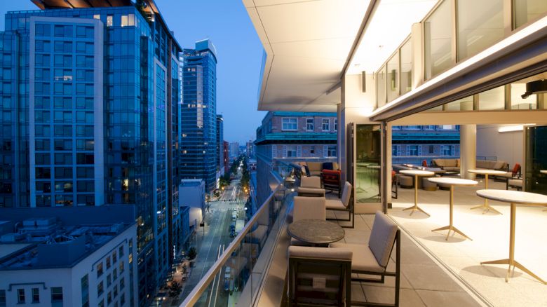 A modern cityscape view at dusk from a high balcony, with seating areas and glass buildings in the background.