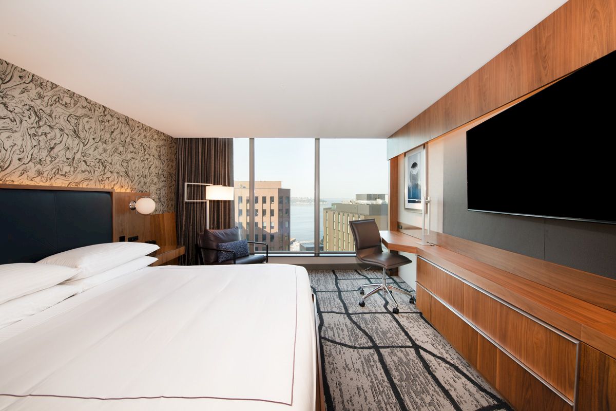 A modern hotel room features a king-sized bed, desk, chair, large TV, and a window with a city view, blending comfort with style.