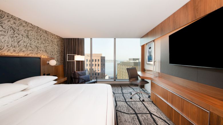 A modern hotel room features a king-sized bed, desk, chair, large TV, and a window with a city view, blending comfort with style.