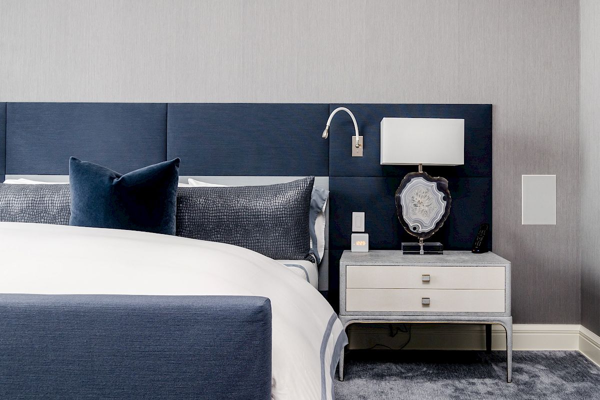 A modern bedroom with a navy blue and white color scheme, featuring a bed, headboard, nightstand, lamp, decorative items, and wall-mounted reading light.