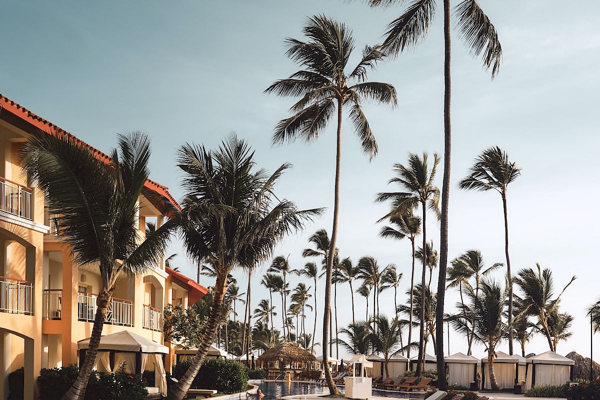 This image shows a tropical resort with a pool, lounge chairs, and palm trees under a clear sky, evoking a relaxing vacation atmosphere.